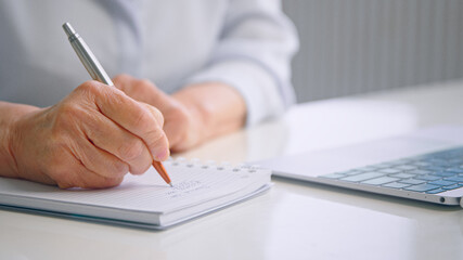 Old lady hand holds grey brown pen and writes notes on paper notebook page near grey laptop on white office table