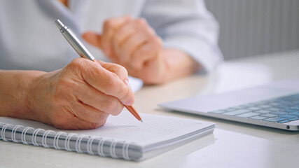 Senior lady teacher with wrinkly hands holds coloured pen over white page of paper notebook near...