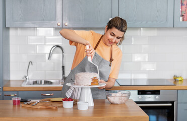 culinary, baking and cooking food concept - happy smiling young woman making layer cake and spreading topping cream on kitchen at home - obrazy, fototapety, plakaty