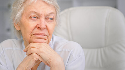 Thoughtful grey haired senior lady looks into window holding head over hands and sitting on white chair in office