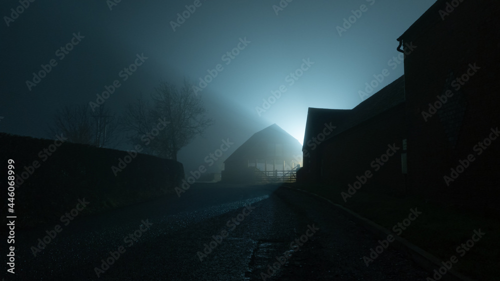 Poster a dark moody country road going into the distance, next to farm buildings back lighted on a foggy wi