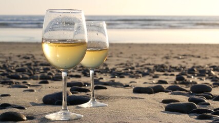 Two wineglasses on sandy ocean beach. Couple of glasses and pebbles, white wine for romantic date near sea water. Cozy beachfront sunset near tide waves. Seamless looped cinemagraph. California, USA.