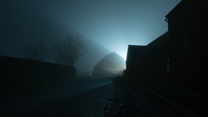 A dark moody country road going into the distance, next to farm buildings back lighted on a foggy winters night. © Dave