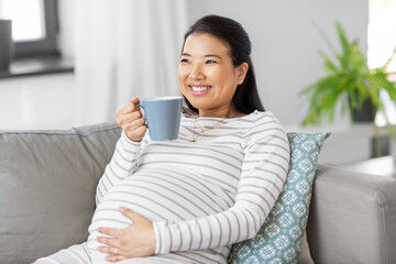 pregnancy, rest, people and expectation concept - happy smiling pregnant asian woman with mug sitting on sofa at home and drinking tea