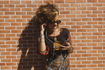 Beautiful African American woman leaning against orange bricks wall wearing earphones listening to music on smartphone and smiling. Copy space for text.