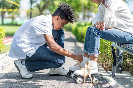 An Asian woman wears roller skates and an African American male ties the shoelaces for her girlfriend to prepare for a activity in the park. 