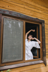Fototapeta premium A beautiful, sleepy brunette bride in white lingerie with a long veil stands in the window against the background of the wall of a wooden house in the morning. Wedding photography.