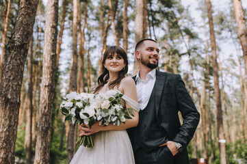 Wedding portrait of the newlyweds. A bearded, stylish groom in a suit and a beautiful brunette bride in a white dress with a bouquet in her hands are standing and hugging in nature in the forest.