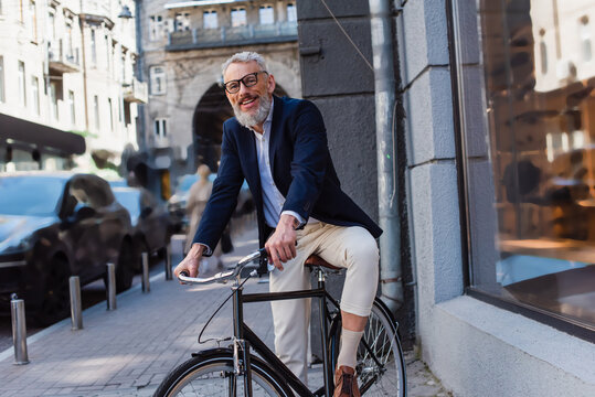 positive and bearded man in glasses riding bicycle on urban street