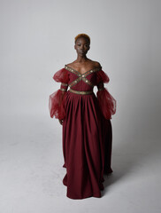 Full length portrait of pretty African woman wearing long red renaissance, medieval fantasy gown, standing pose on a light grey studio background.