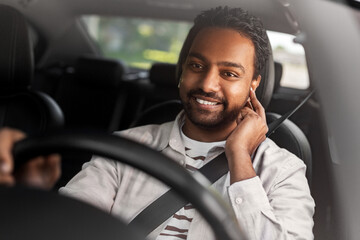 Obraz premium transport, people and technology concept - smiling indian man or driver with wireless earphones or hands free device driving car