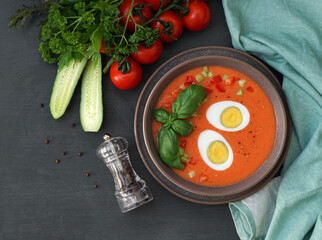 delicious gazpacho soup with eggs, basil and ingredients next to a plate on a gray background with a mintcolor napkin. Place for text. top view      
