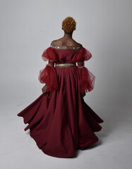 Full length portrait of pretty African woman wearing long red renaissance medieval fantasy gown, standing pose on a light grey studio background.