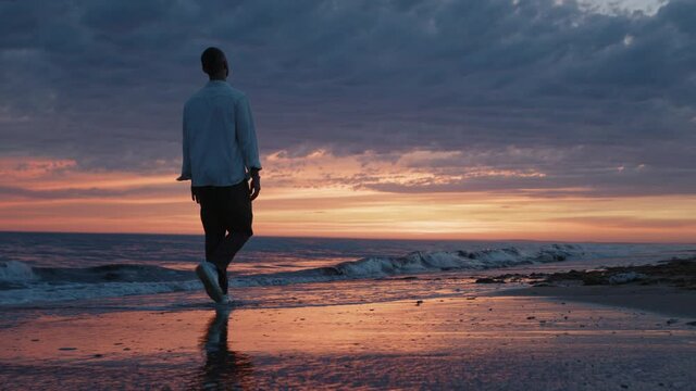 Handsome happy young hipster man slowly walks alone along beach at the end of a day horizon during sunset. Attractive romantic men traveler lifestyle. Freedome peaceful people concept slow motion.