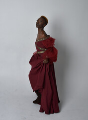 Full length portrait of pretty African woman wearing long red renaissance medieval fantasy gown, standing pose on a light grey studio background.