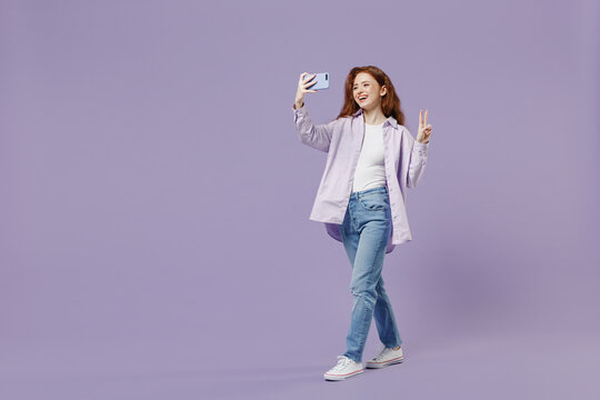Full size body length young redhead woman 20s wears white T-shirt violet jacket do selfie shot on mobile cell phone show victory sign isolated on pastel purple color wall background studio portrait.