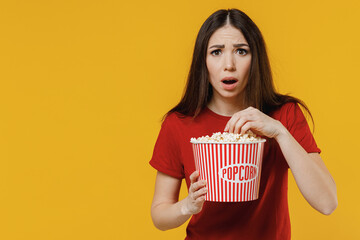 Scared frightened shocked scared young brunette woman 20s wears basic red t-shirt watch movie film holding bucket of popcorn eat keeping mouth wide open isolated on yellow background studio portrait.