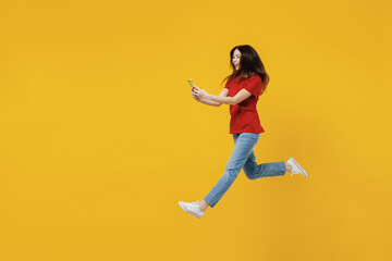Fototapeta na wymiar Full size body length excited young brunette woman 20s wears basic red t-shirt jump hold in hand use mobile cell phone isolated on yellow background studio portrait. People emotions lifestyle concept.