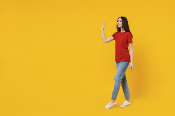 Fototapeta na wymiar Full size body length side view profile young brunette woman 20s wear basic red t-shirt stand go move step meet greet waving hand isolated on yellow background studio portrait. People emotion concept