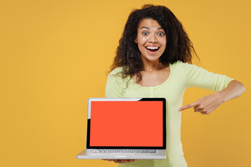 Amazed shocked african american young brunette woman 20s wears green shirt hold use point on work on laptop pc computer with blank screen workspace area isolated on yellow background studio portrait.