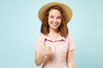 Smiling cheery young redhead curly woman 20s showing thumb up wears casual pink dress straw hat look camera isolated on pastel blue color background studio portrait. People emotions lifestyle concept