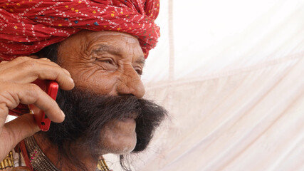 Portrait of an Indian old male with a red turban and mustache talking on a phone with a happy face