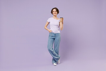 Full length young smiling satisfied fun happy woman 20s in white t-shirt hold takeaway delivery craft paper brown cup coffee to go stand akimbo isolated on pastel purple background studio portrait