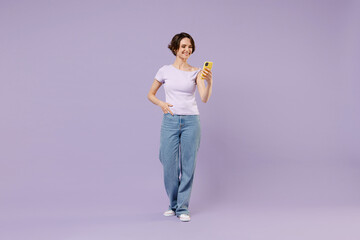 Full length young smiling happy brunette woman 20s with bob haircut in white t-shirt using mobile cell phone chat online browsing surfing internet isolated on pastel purple background studio portrait