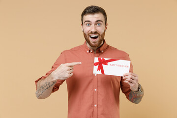 Shocked amazed tatooed young brunet man 20s with short haircut wears apricot shirt hold pointing on gift certificate coupon voucher card for store isolated on pastel orange background studio portrait
