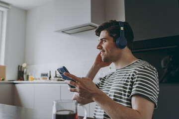 Young side view smiling happy man in striped t-shirt headphones using mobile cell phone look aside listen to music browsing sitting by table in light kitchen at home alone. People lifestyle concept