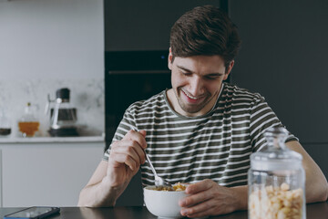 Close up young man 20s in casual clothes striped t-shirt eat breakfast muesli cereals with milk fruit in bowl sit at table cooking food in light kitchen at home alone. Healthy diet lifestyle concept.