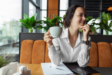 Smiling businesswoman in formal wear drinking coffee in cafe while having coffee break during remote work and looking away 