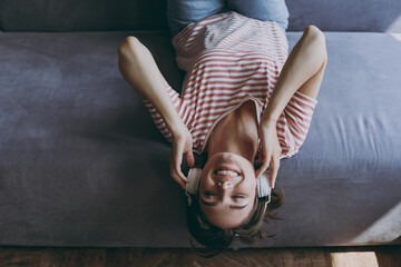 Top view from above young woman in casual clothes headphones lying on grey sofa listen to music new playlist enjoy free time rest on weekends indoors flat at home. People lifestyle leisure concept