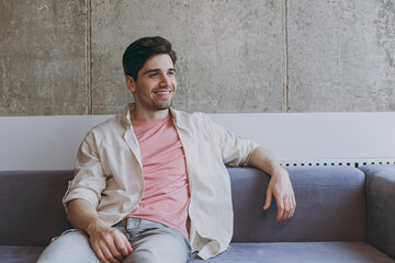 Young fun smiling happy minded caucasian man 20s wearing casual clothes beige shirt pink t-shirt look aside sitting on grey sofa resting indoors at home on weekends. People lifestyle leisure concept
