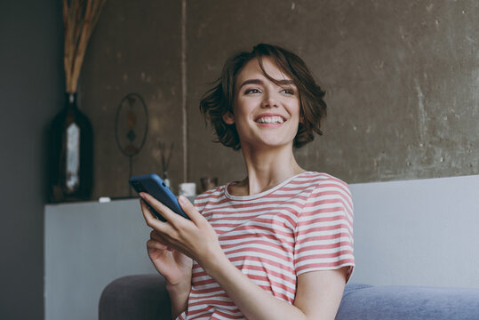 Young wistful happy caucasian smiling woman 20s in casual clothes sitting on grey sofa using mobile cell phone chat online look aside rest relax indoors flat at home People lifestyle leisure concept. - Powered by Adobe