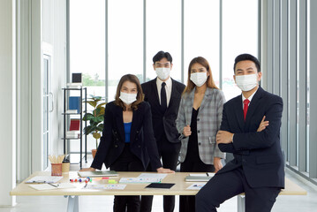 Asian businessman in a suit leaning against the table with a confident expression. The team is preparing for the meeting in the background. Everyone wears a mask. Concept of leadership.