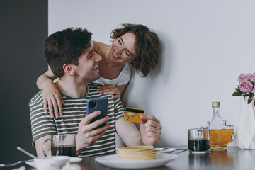 Young cheerful smiling couple two woman man in casual clothes sit by table eat pancake use mobile cell phone credit bank card shop online order delivery in light kitchen at home. Healthy diet concept