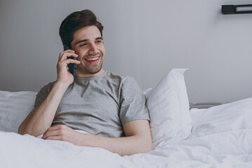Calm satisfied smiling happy young man 20s in pajamas grey t-shirt lying sit in bed talking by...