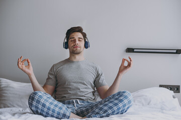 Full body young man in blue pajamas headphones lying sit in bed hold spread hands in yoga om aum...