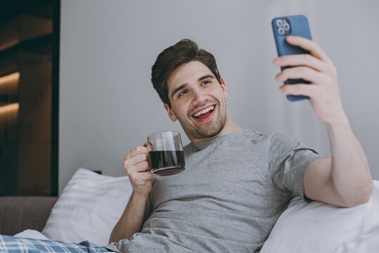 Cheerful young man wear pajamas grey t-shirt lying sit in bed hold cup drink coffee do selfie shot on modile cell phone resting relax at home indoors bedroom Good mood night morning bedtime concept