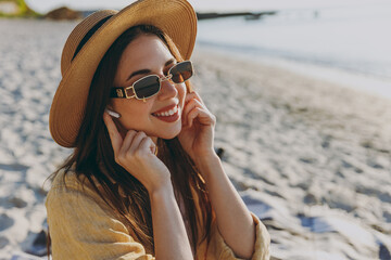 Young sunlit happy traveler tourist woman wear straw hat glasses shirt summer clothes wireless earphones listen to music outdoors on sea beach background. People vacation lifestyle journey concept