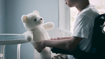 Disabled child on wheelchair talking with a white teddy bear in hands, emotional concept, Lifestyle of disability kid home alone, the best friend is toy. - obrazy, fototapety, plakaty
