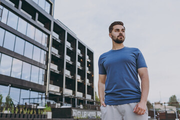 Fototapeta premium Bottom view young european strong sporty athletic toned fit sportsman man 20s wearing blue casual t-shirt walking strolling going looking aside in summer cloudy day morning near hotel black building.