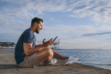 Full size side view young strong sporty fit sportsman man in sports clothes shorts warm up train chat by mobile cell phone at sunrise sun over sea beach outdoor on pier seaside in summer day morning.