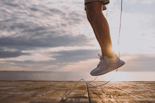 Cropped up photo shot young sporty fit sportsman man in sports clothes shorts warm up training jump on skipping rope at sunrise sun dawn over sea beach outdoor on pier seaside in summer day morning - Powered by Adobe