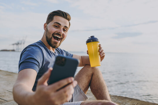 Young cool sporty strong sportsman man in sports clothes shorts train do selfie shot photo on mobile cell phone drink water at sunrise sun over sea beach outdoor on pier seaside in summer day morning.