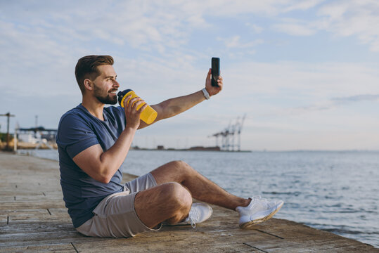 Full body young sporty sportsman man in sports clothes shorts train do selfie shot photo on mobile cell phone drink water at sunrise sun over sea beach outdoor on pier seaside in summer day morning.