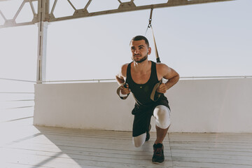 Full size young strong sporty athletic toned fit sportsman man in sports clothes warm up suspended training with TRX work out at sunrise sun dawn over sea beach seaside outdoor in summer day morning
