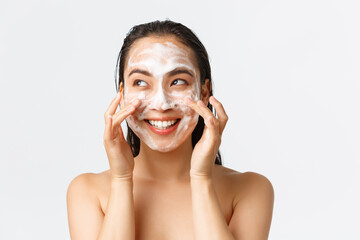 Skincare, women beauty, hygiene and personal care concept. Happy cheerful pretty asian girl standing naked in bathroom and apply cleansing foam on face, smiling and looking left - obrazy, fototapety, plakaty