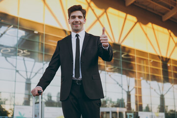 Bottom view young traveler brunet businessman man in black classic suit stand outside at international airport terminal with suitcase valise show thumb up. Air flight business trip lifestyle concept.